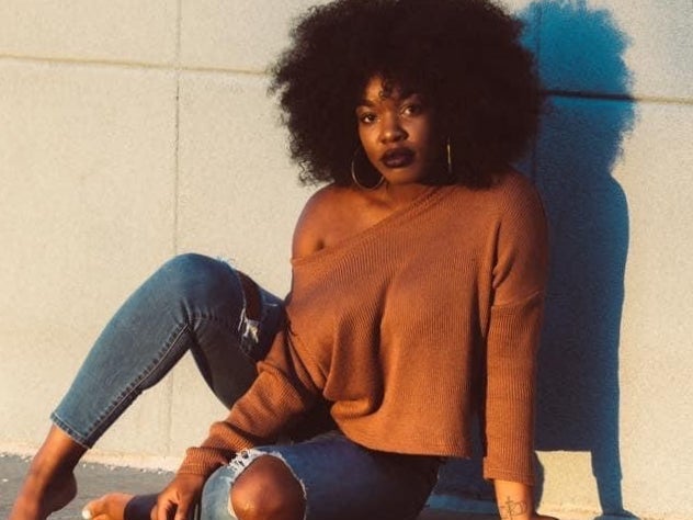 Woman with an afro in a rust sweater seated against a sunlit wall.